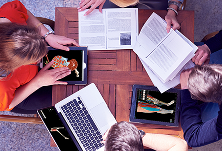 4 Studierende lernen an einem Tisch mit geöffneten Lehrbüchern, 2 Tablets und einem Laptop, auf deren Bildschirmen Visible Body 3D-Anatomie angezeigt wird
